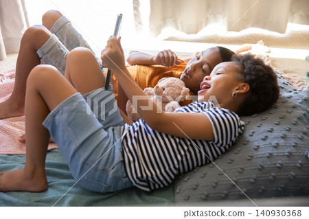 Diverse siblings holding tablet above heads and hugging teddy bear while lying on bed near window Diverse siblings holding tablet above heads and hugging teddy bear while lying on bed near window 140930368