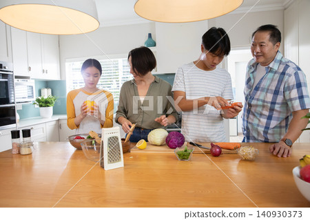 Diverse family slicing carrots and oranges on wooden island in home kitchen with glass bowl 140930373