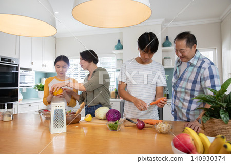 Diverse family of four standing around kitchen island slicing bell pepper and peeling carrot 140930374