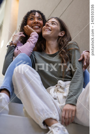 Diverse female friends hugging while sitting on stair steps in stairwell under light fixture Diverse female friends hugging while sitting on stair steps in stairwell under light fixture 140930386