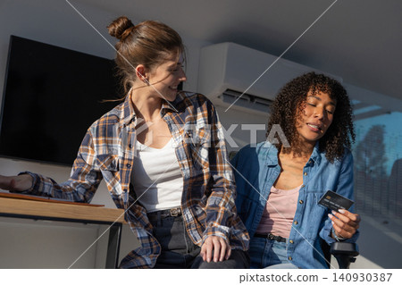 Diverse female coworkers discussing payment while holding credit card at office desk Diverse female coworkers discussing payment while holding credit card at office desk 140930387