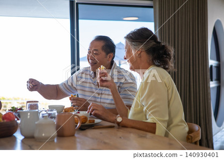 Diverse senior couple sharing breakfast at wooden dining table with fruit basket and mugs Diverse senior couple sharing breakfast at wooden dining table with fruit basket and mugs 140930401