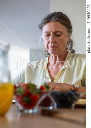 Senior woman sitting at wooden table in home kitchen chopping berries into glass bowls Senior woman sitting at wooden table in home kitchen chopping berries into glass bowls 140930403