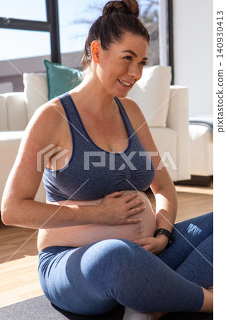 Pregnant woman sitting cross-legged on yoga mat in living room, cradling belly and smiling Pregnant woman sitting cross-legged on yoga mat in living room, cradling belly and smiling 140930413