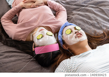 Asian mother and daughter resting on bed wearing facial masks, cucumber slices on eyes Asian mother and daughter resting on bed wearing facial masks, cucumber slices on eyes 140930423