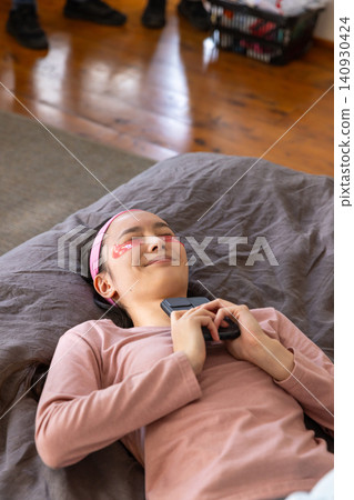 Asian woman lying at home on gray bedding wearing pink headband with gel patches holding mobile 140930424
