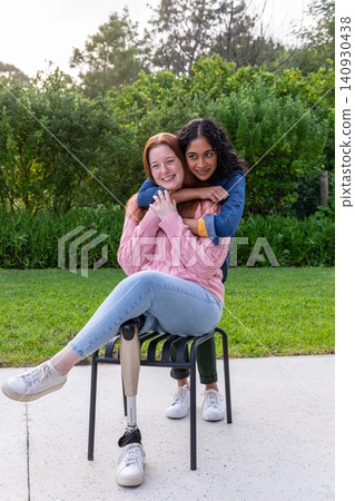 Diverse women embracing on metal chair on concrete patio in garden with prosthetic leg, sneakers Diverse women embracing on metal chair on concrete patio in garden with prosthetic leg, sneakers 140930438