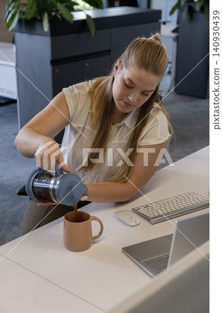 White woman in twenties pouring coffee from french press into mug at desk with laptop White woman in twenties pouring coffee from french press into mug at desk with laptop 140930439