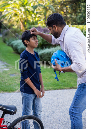 Indian father with son adjusting blue bicycle helmet strap beside bicycle on paved garden path Indian father with son adjusting blue bicycle helmet strap beside bicycle on paved garden path 140930452