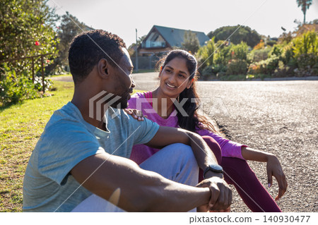 Diverse couple sitting on suburban street, resting after run, checking fitness watch Diverse couple sitting on suburban street, resting after run, checking fitness watch 140930477