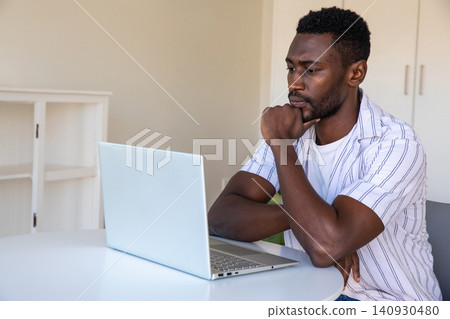 African american man resting chin on hand in home office, gazing at laptop, copy space African american man resting chin on hand in home office, gazing at laptop, copy space 140930480