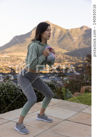 Asian korean woman performing weighted squat in sportswear on tiled terrace holding kettlebell Asian korean woman performing weighted squat in sportswear on tiled terrace holding kettlebell 140930516