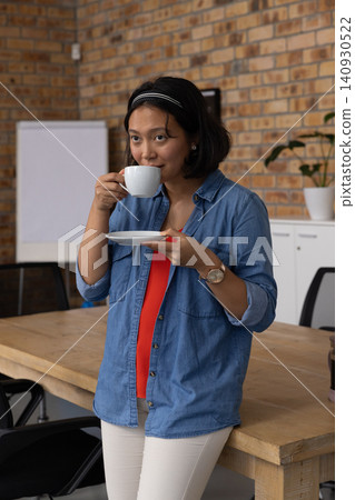 Asian woman sipping coffee from cup beside conference table in meeting room with flip chart Asian woman sipping coffee from cup beside conference table in meeting room with flip chart 140930522