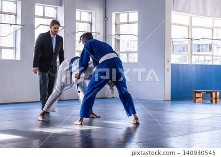Diverse male athletes wearing gis grappling under referee watch in dojo on blue mats near bench 140930545