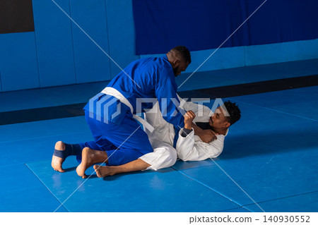 African american men grappling on blue mats in martial gym wearing gis with white belt African american men grappling on blue mats in martial gym wearing gis with white belt 140930552