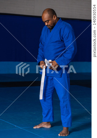 Senior african american man tying white belt on gi standing barefoot on mats at dojo 140930554