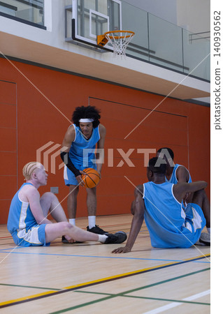 Diverse male teammates wearing jerseys holding basketball under hoop and sitting on gym court Diverse male teammates wearing jerseys holding basketball under hoop and sitting on gym court 140930562