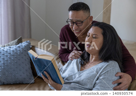 Diverse couple sitting on tan leather sofa in living room reading hardcover book with eyeglasses Diverse couple sitting on tan leather sofa in living room reading hardcover book with eyeglasses 140930574