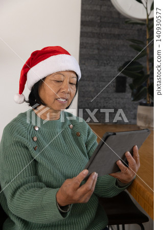 Senior Asian woman sitting at table holding tablet wearing Santa hat, green sweater, copy space Senior Asian woman sitting at table holding tablet wearing Santa hat, green sweater, copy space 140930577