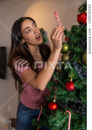 Woman in pink shirt, jeans hanging candy cane ornament on christmas tree in living room Woman in pink shirt, jeans hanging candy cane ornament on christmas tree in living room 140930582
