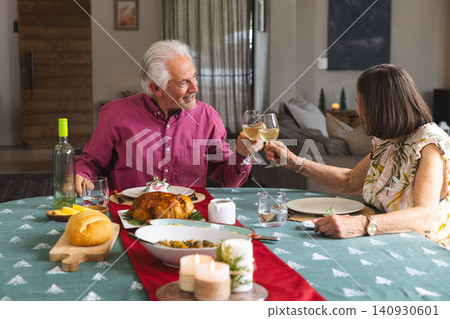 Senior couple raising wine glasses and toasting at home dining table with roast chicken, salad bowl Senior couple raising wine glasses and toasting at home dining table with roast chicken, salad bowl 140930601
