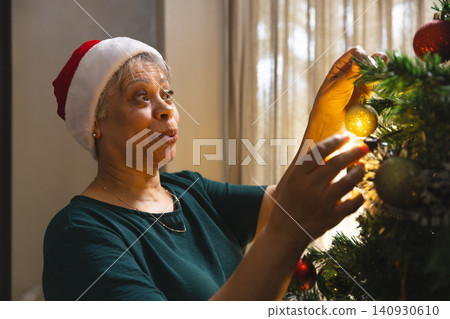 Senior African American wearing Santa hat adding gold ornament to Xmas tree at home, copy space Senior African American wearing Santa hat adding gold ornament to Xmas tree at home, copy space 140930610