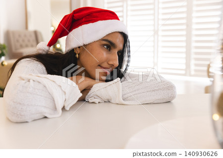 Woman leaning on tabletop in living room wearing knit sweater with Santa hat near Christmas tree Woman leaning on tabletop in living room wearing knit sweater with Santa hat near Christmas tree 140930626