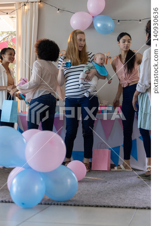 Diverse female friends gathering around home table holding infant among balloons, gift bags Diverse female friends gathering around home table holding infant among balloons, gift bags 140930636