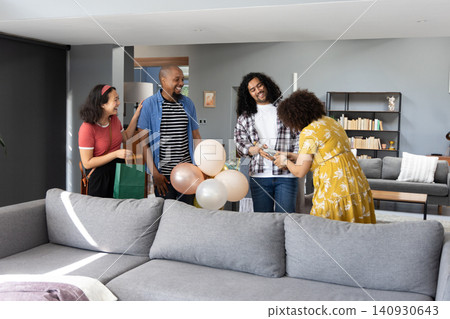 Diverse friends gathering in living room at sofa, handing wrapped gift, holding pastel balloons Diverse friends gathering in living room at sofa, handing wrapped gift, holding pastel balloons 140930643