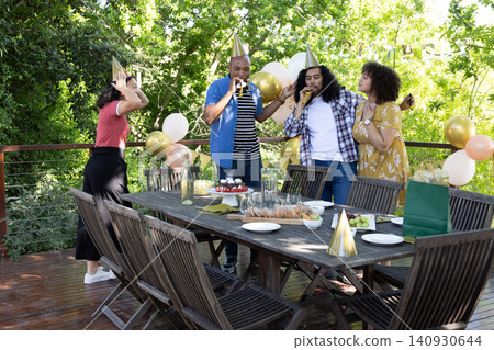 Diverse friends wearing party hats blowing noisemakers around table on deck with cake, balloons Diverse friends wearing party hats blowing noisemakers around table on deck with cake, balloons 140930644