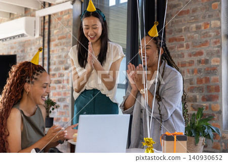 Diverse female coworkers in party hats clapping and smiling around laptop with gift box at office Diverse female coworkers in party hats clapping and smiling around laptop with gift box at office 140930652
