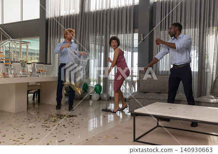 Diverse friends cleaning up metallic confetti in modern living room with broom, trash bag Diverse friends cleaning up metallic confetti in modern living room with broom, trash bag 140930663