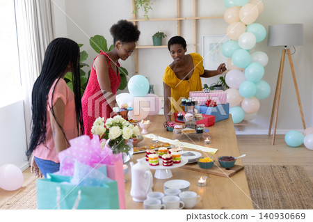 African American friends preparing cupcakes on table under pastel balloons in bright dining room African American friends preparing cupcakes on table under pastel balloons in bright dining room 140930669
