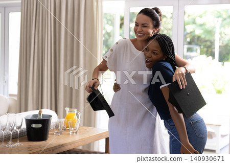 Mother and daughter embracing and holding graduation caps in dining area by table with wine glasses 140930671