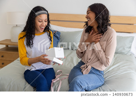 Diverse mom and teenage daughter opening white gift box on bed in bedroom with pink ribbon bag 140930673