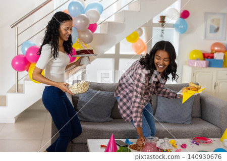 Diverse mom and teenage daughter carrying cake with candles and popcorn toward table Diverse mom and teenage daughter carrying cake with candles and popcorn toward table 140930676