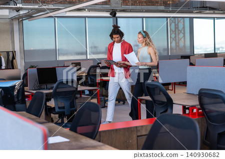 Diverse coworkers walking while consulting tablet and printed documents through open-plan office Diverse coworkers walking while consulting tablet and printed documents through open-plan office 140930682