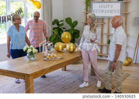 Diverse senior friends standing around wooden dining table in living room admiring balloons Diverse senior friends standing around wooden dining table in living room admiring balloons 140930685