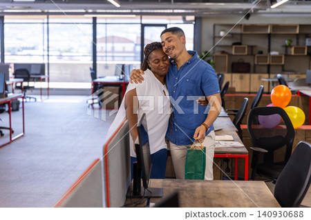 Diverse coworkers hugging beside desk in modern workspace with gift bag and balloons Diverse coworkers hugging beside desk in modern workspace with gift bag and balloons 140930688
