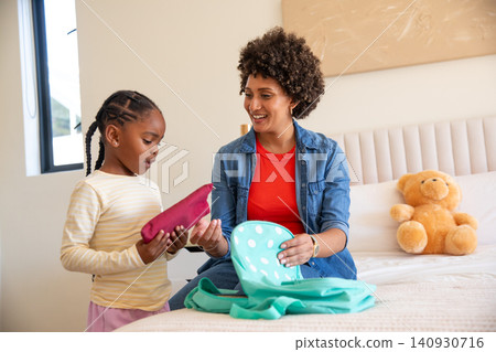 Diverse mother and daughter packing blue polka dot backpack on bed in bedroom next to teddy bear Diverse mother and daughter packing blue polka dot backpack on bed in bedroom next to teddy bear 140930716