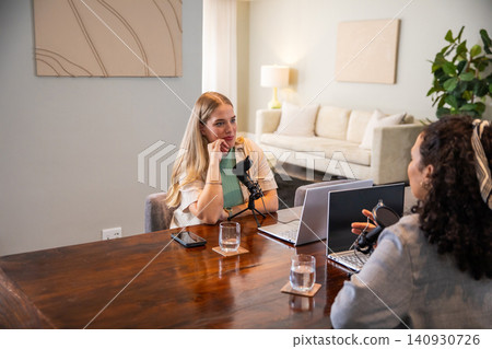 Diverse female coworkers sitting at table at home co-hosting podcast with microphones, laptops Diverse female coworkers sitting at table at home co-hosting podcast with microphones, laptops 140930726