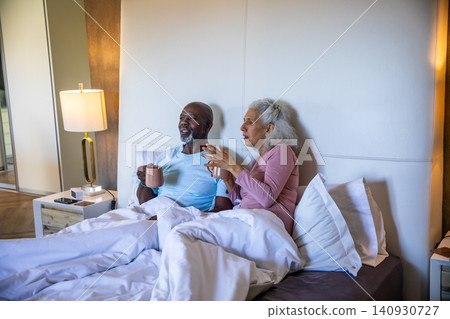 Diverse senior couple sitting on bed in bedroom sipping from mugs with smartphone and glasses Diverse senior couple sitting on bed in bedroom sipping from mugs with smartphone and glasses 140930727