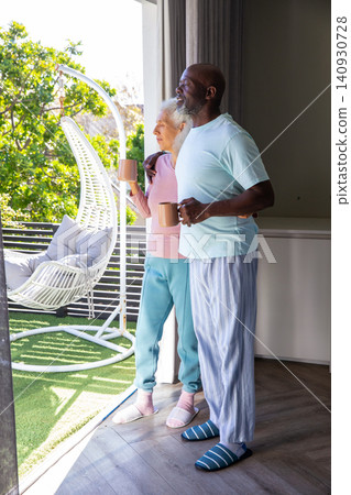 Senior couple standing at door at home, holding ceramic mugs, wearing slippers, gazing onto balcony Senior couple standing at door at home, holding ceramic mugs, wearing slippers, gazing onto balcony 140930728