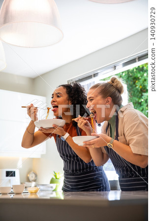 Diverse female friends tasting noodles in bowls with forks at kitchen island in striped aprons Diverse female friends tasting noodles in bowls with forks at kitchen island in striped aprons 140930729