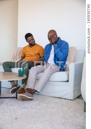 African american father and son sitting on sofa at home holding smartphone, coffee mug African american father and son sitting on sofa at home holding smartphone, coffee mug 140930746