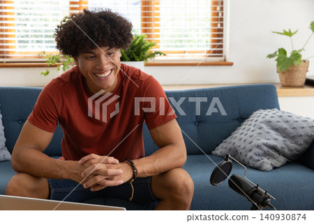 African american man sitting on blue sofa in living room using pop filter microphone, laptop African american man sitting on blue sofa in living room using pop filter microphone, laptop 140930874