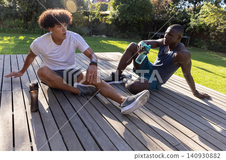 Diverse male friends resting on wooden deck in backyard in sportswear and holding bottles Diverse male friends resting on wooden deck in backyard in sportswear and holding bottles 140930882