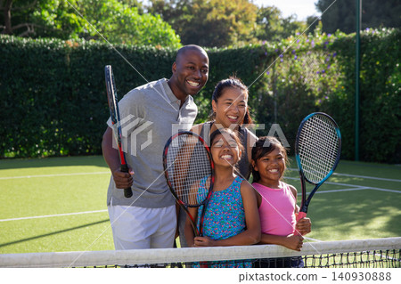 Diverse family standing behind tennis net on green turf court holding tennis rackets smiling Diverse family standing behind tennis net on green turf court holding tennis rackets smiling 140930888