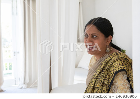 Mature indian woman standing between bed and balcony wearing gold sari and jhumkas, copy space Mature indian woman standing between bed and balcony wearing gold sari and jhumkas, copy space 140930900