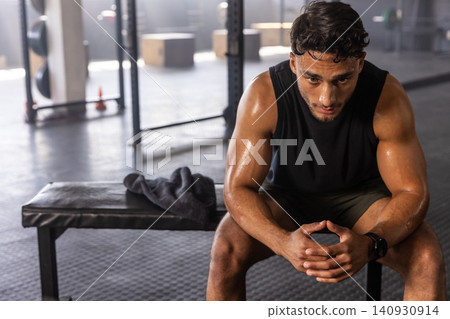 Man sitting on padded bench sweating in weight area wearing tank top, squat rack, copy space Man sitting on padded bench sweating in weight area wearing tank top, squat rack, copy space 140930914